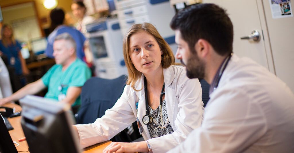 Two young medical students in white coats discussing a patient case at a hospital workstation.