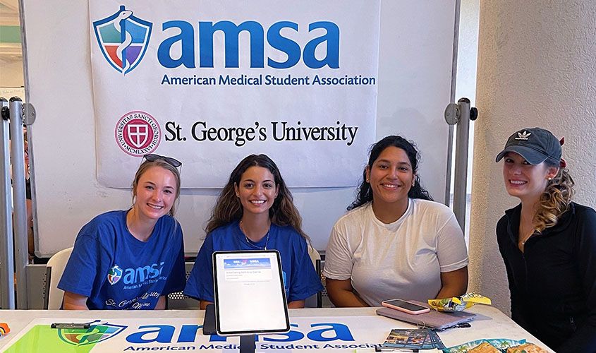 Four St. George's University (SGU) medical students staffing an AMSA (American Medical Student Association) booth during a campus club fair. News