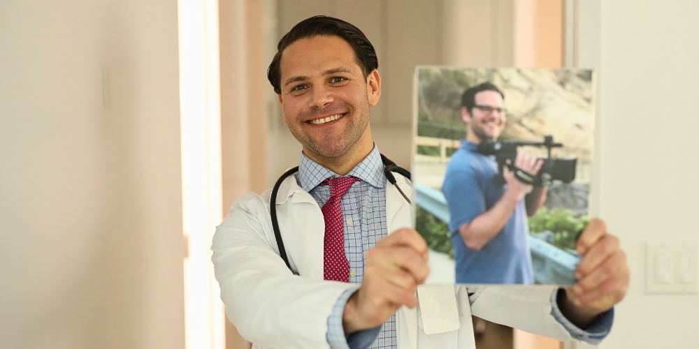 Smiling physician in a white coat and stethoscope holding up a photo of his younger self holding a video camera