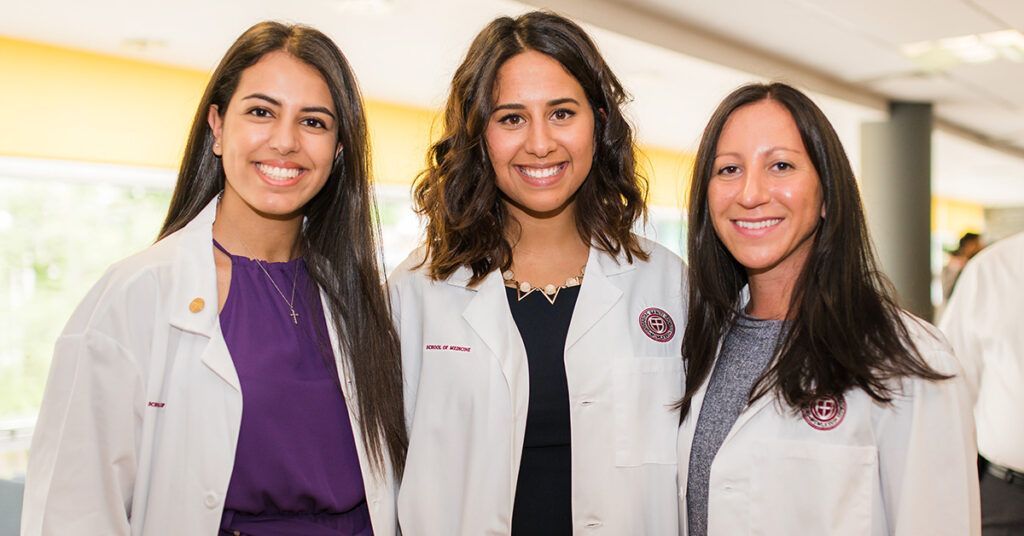 Three female medical students or physicians in white lab coats smiling during an event.