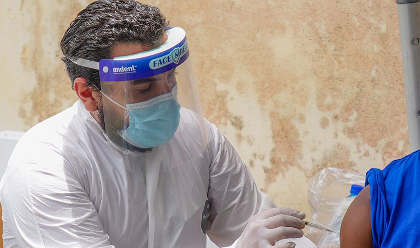 A healthcare worker in full white PPE and a face shield administers a vaccine via injection at a community clinic. News