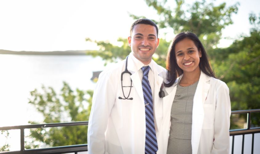 Two St. George's University (SGU) medical students or physicians in white coats and stethoscopes, smiling outdoors. News