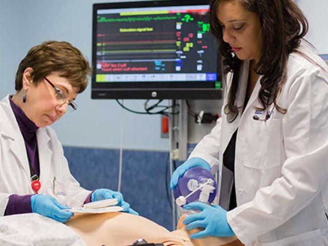 Two SGU medical students wearing lab coats and gloves working with a manikin and monitoring equipment.
